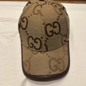 Beige keps från Gucci - Snygg beige keps från Gucci med brunt GG-mönster. Kepsen har en justerbar rem baktill och är perfekt för en stilren look. Tillverkad i ett slitstarkt material med en klassisk design.