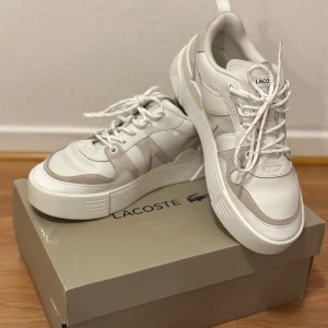 Vita sneakers från Lacoste - Snygga vita sneakers från Lacoste med stilren design och snörning. Skorna har en kombination av skinn och mocka, vilket ger en modern look. Perfekta för en avslappnad stil.