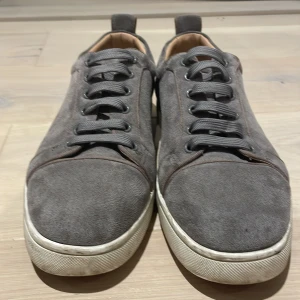 Grå sneakers från Christian Louboutin - Snygga grå sneakers från Christian Louboutin med klassisk röd sula. Skorna har snörning och är tillverkade i mocka med en bekväm insida i skinn. Perfekta för en stilren look.