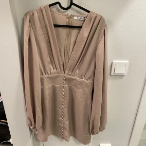 Champagnefärgad satin klänning från NA-KD - Elegant champagnefärgad klänning i satin från NA-KD med knappar framtill och djup v-ringning. Klänningen har långa ärmar och en dragkedja i ryggen. Perfekt för en stilfull look.