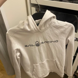 Vit hoodie från Sail Racing - Säljer en stilren vit hoodie från Sail Racing med logga på bröstet. Tröjan har en klassisk känguruficka och en bekväm huva. Perfekt för en avslappnad stil. Produkten är i bra skick och är i storlek 160