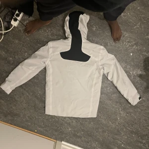 Vit och svart jacka från Sail Racing - Snygg vit och svart jacka från Sail Racing med huva och dragkedja. Jackan har flera praktiska fickor och en stilren design med svarta detaljer på bröstet och ärmarna.