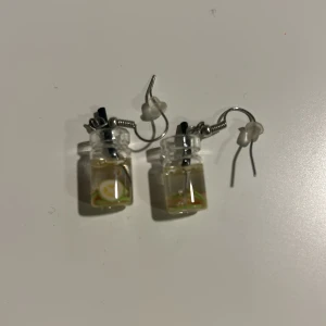 Örhängen med små glasflaskor - Unika örhängen med små glasflaskor fyllda med en klar vätska och en grön detalj inuti. De har en krok för enkel användning och ger en kreativ touch till din stil.Tvättar innan skickar 