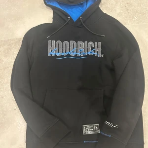 Hoodrich blend hoodie - Säljer en svart hoodie från Hoodrich med blå detaljer och text på bröstet och ryggen. Hoodien har en känguruficka och en justerbar huva med blått foder. Perfekt för en avslappnad stil.