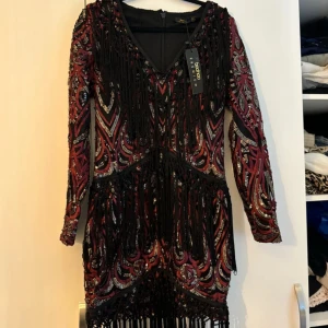 Mönstrad klänning från Boohoo Premium - Snygg klänning från Boohoo Premium med ett färgstarkt mönster i rött och svart. Klänningen har långa ärmar och en v-ringad design med fransdetaljer och paljetter. Perfekt för en festlig kväll!