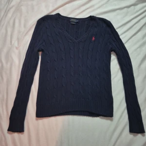 Mörkblå stickad tröja från Ralph Lauren - Säljer en snygg mörkblå stickad tröja från Ralph Lauren i pima bomull. Tröjan har en v-ringning och det klassiska röda Polo-logotypen broderad på bröstet. Perfekt för en stilren look!