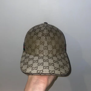 Beige Gucci keps - Snygg beige keps från Gucci med det ikoniska GG-mönstret. Kepsen har en justerbar rem baktill i brunt skinn och är perfekt för en stilren look. Passar bra till både vardag och speciella tillfällen. Pris kan diskuteras vid snabb affär!!