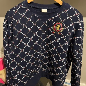 Mönstrad mörkblå tröja - Snygg mörkblå tröja med ett unikt kedjemönster i vitt och rött. Tröjan har ett emblem på bröstet och långa ärmar. Perfekt för en stilren look.