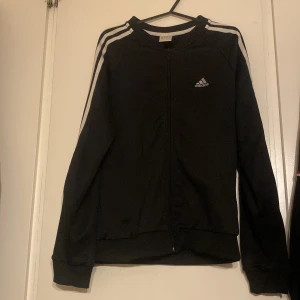 Svart träningsjacka från Adidas - Snygg svart träningsjacka från Adidas med klassiska vita ränder längs ärmarna och dragkedja framtill. Adidas-logga på bröstet och glittrig text på ryggen. Perfekt för sportiga tillfällen.