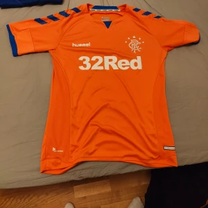 Rangers tredjetröja 18/19 - Säljer en Rangers tröja 2018/2019 från Hummel med blå detaljer och vit text. Tröjan har korta ärmar och är märkt med '32Red' och 'utilita'. Perfekt för fotbollsfans!