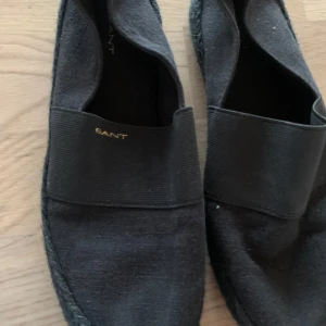 Svarta loafers från Gant - Snygga svarta loafers från Gant med elastisk rem över foten för extra komfort. Skorna har en diskret logga på sidan och en mönstrad sula för bra grepp. Perfekta för en stilren look.storlek 38 . Den har använt bara två gånger 