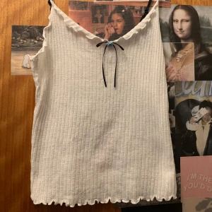 Vit linnetopp med rosett Zara - Söt vit linnetopp med smala axelband och en liten blå ros och svart rosett framtill. Toppen har en vågig kantdetalj vid halsringningen och nederkanten, vilket ger en romantisk touch. Perfekt för varma dagar eller som ett lager under en kofta.