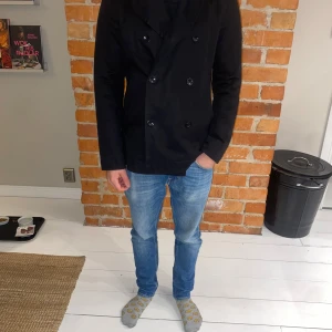 Svart skepparkavaj från Filippa K - Snygg svart skepparkavaj från Filippa K i herrstorlek S. Jackan har en klassisk dubbelknäppning med svarta knappar och är perfekt för både höst och vår. Den har långa ärmar och en stilren design som passar till många olika outfits.