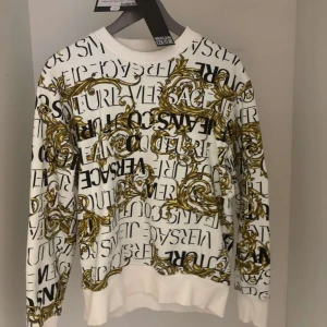 Versace Jeans Couture tröja - Snygg vit tröja från Versace Jeans Couture med ett iögonfallande mönster i svart och guld. Tröjan har långa ärmar och en rund halsringning. Perfekt för dig som vill sticka ut med stil!