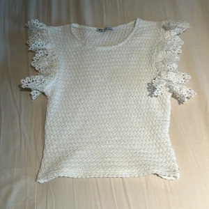 Vit spetsdetaljerad topp från Zara - Säljer en söt vit topp från Zara med vackra spetsdetaljer på ärmarna. Toppen har en fin textur och är perfekt för en somrig look.