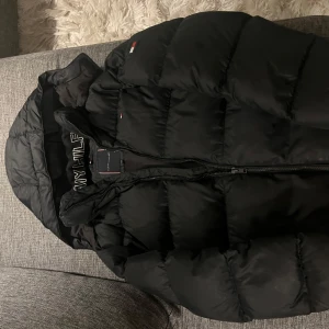 Svart pufferjacka från Tommy Hilfiger - Snygg svart pufferjacka från Tommy Hilfiger med dragkedja och huva. Perfekt för kyliga dagar med sin vadderade design som håller dig varm. Använd fåtal gånger.