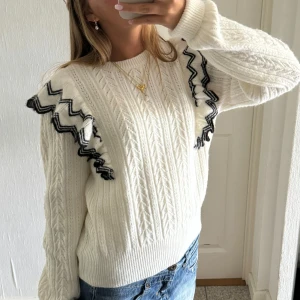 Vit stickad tröja med volangdetaljer - Så söt!!!! stilren vit stickad tröja med svarta volangdetaljer på axlarna. Tröjan har ett klassiskt flätmönster och långa ärmar. Ej använd mycket så i jättebra skick!! Strl 170 men sitter som en s-m