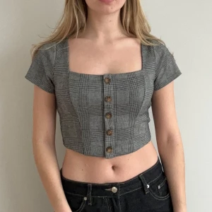 Grå rutig croppad topp - Säljer en stilren grå rutig croppad topp med korta ärmar och knappar framtill. Perfekt för en trendig look!