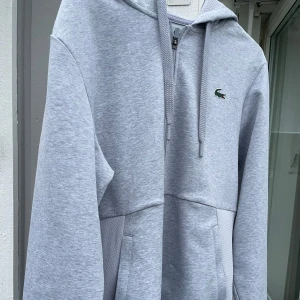 Grå hoodie från Lacoste helt ny är för stor för mig som är 170cm  - Säljer en stilren grå hoodie från Lacoste med dragkedja och klassisk krokodil-logga på bröstet. Tröjan har en bekväm passform och är perfekt för en avslappnad stil. Den är tillverkad i mjukt material och har en praktisk ficka framtill.