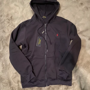 Marinblå hoodie från Polo Ralph Lauren - Säljer en stilren marinblå hoodie från Polo Ralph Lauren med dragkedja och det klassiska röda broderade logotypen på bröstet. Perfekt för en avslappnad look. Hoodien har långa ärmar och en bekväm passform.