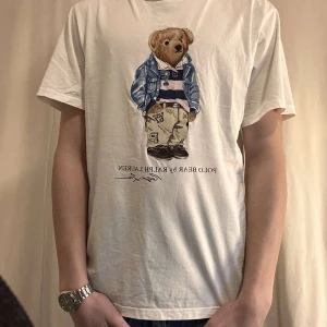 Vit t-shirt med Polo Bear från Ralph Lauren - Vit t-shirt från Ralph Lauren med det ikoniska Polo Bear-trycket på framsidan. T-shirten har en klassisk passform och korta ärmar, perfekt för en avslappnad stil.
