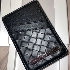Svart flätad kortplånbok från iDeal of Sweden - Säljer en stilren svart kortplånbok från iDeal of Sweden med flätad design. Perfekt för att hålla dina kort säkra och organiserade. Den är magnetisk och smidig att använda. Helt ny aldrig använd där original förpackning ingår.
