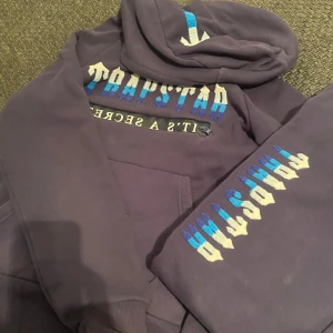 Mörkblå hoodie med textdetaljer - Säljer en mörkblå hoodie med coola textdetaljer i blått och vitt. Hoodien har en stor ficka framtill och en bekväm huva. Perfekt för en avslappnad stil.