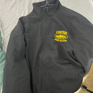 Svart jacka från Cortez - Säljer en svart jacka från Cortez med gul logga på bröstet och text på kragen. Jackan har en dragkedja framtill och hög krage. Perfekt för en cool och avslappnad stil.