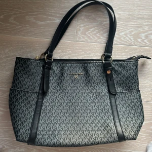 Svart och grå axelväska från Michael Kors - Snygg axelväska från Michael Kors i svart och grått med mönstrat tyg och guldfärgade detaljer. Väskan har dubbla handtag och en dragkedja upptill. Perfekt för att bära dina dagliga nödvändigheter med stil. Bra skick och kvalitet. Den har dock två defekter vilket syns på sista bilden. dels har en av knapparna på framsidan låtsnat men vi har lagat det så att det ej syns så mycket och sen är den lite fläckig på insidan.