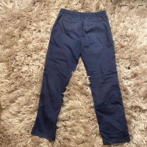 Blå byxor från Polo Ralph Lauren - Snygga blå byxor från Polo Ralph Lauren med elastisk midja och knappstängning. Byxorna har två bakfickor med knappar och en liten broderad logga i rött. Perfekta för en stilren look.