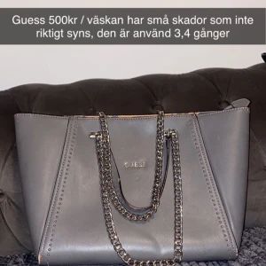 Grå axelväska från Guess - Snygg grå axelväska från Guess med kedjedetaljer och dragkedja. Väskan har ett rymligt inre med logotypmönster och flera fack. Perfekt för att bära med sig det nödvändigaste med stil.