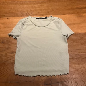 Ljusgrön ribbad topp från Vero Moda - Säljer en ljusgrön ribbad topp från Vero Moda. Toppen har korta ärmar och en rund halsringning med vågiga kanter vid ärmslut och nederkant. Perfekt för en avslappnad stil.