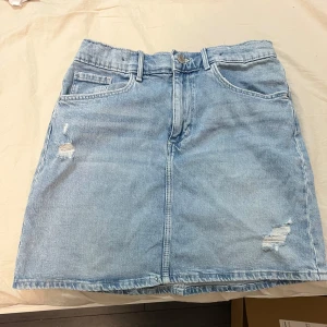 Blå jeanskjol från hm - Säljer en snygg blå jeanskjol från hm. Kjolen har en klassisk design med slitna detaljer och femficksmodell. Perfekt för en avslappnad stil. Den har en knapp och dragkedja framtill för enkel påklädning.