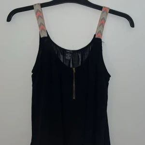 Svart linne med mönstrade axelband - Snyggt svart linne från Rue21 med färgglada mönstrade axelband och dragkedja framtill. Lite genomskinligare tygg. Perfekt för en mer bekväm men endå fin stil 