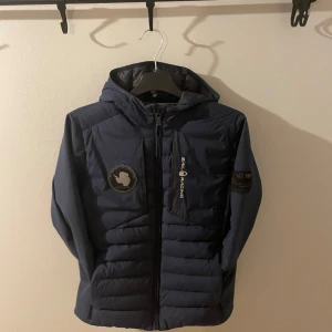 Mörkblå jacka från Sail Racing - Säljer en mörkblå jacka från Sail Racing med luva och drag kedja med en patch på ärmen och en logga på bröstet. Perfekt för kyliga dagar med sin pufferdesign.