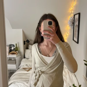 Beige omlottopp med knyt - Snygg beige omlottopp med knyt i midjan. Toppen har långa ärmar och en stilren design som passar perfekt till jeans eller kjol. Perfekt för en avslappnad men chic look. Från Gina storlek xs. 