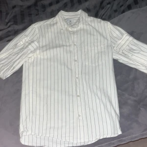 Vit och svart randig skjorta - Snygg vit skjorta med svarta ränder. Skjortan har långa ärmar och knappar framtill. Perfekt för en stilren look.använd en gång och är i ny skick