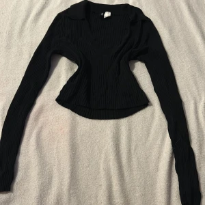 Svart ribbad tröja från Divided - Säljer en svart ribbad tröja från Divided med v-ringning och långa ärmar. Perfekt för en stilren look. Tröjan har en klassisk krage och är i en bekväm passform. Passar bra till både jeans och kjol.