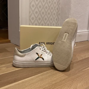 Vita sneakers från Axel Arigato - Snygga vita sneakers från Axel Arigato med en broderad fågel på sidan. Skorna har snörning och grå detaljer på tån. Perfekta för en stilren look. Använda ett fåtal par gånger bara, så riktigt fräscha!