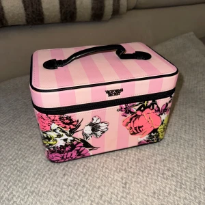 Rosa randig makeup väska från Victoria's Secret - Säljer en superfin rosa randig makeup väska från Victoria's Secret med blommigt mönster. Väskan har ett praktiskt handtag och dragkedja. Perfekt för att organisera dina saker med stil. Insidan är svart och randig med flera fack.