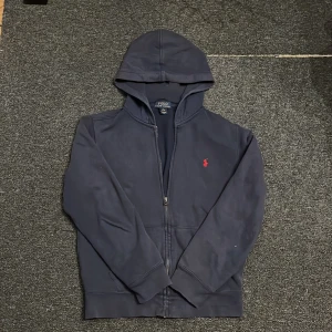 Mörkblå hoodie från Polo Ralph Lauren - Säljer en snygg mörkblå hoodie från Polo Ralph Lauren med dragkedja och det klassiska röda logotypen på bröstet. Perfekt för en avslappnad stil. Den har långa ärmar och en praktisk huva.