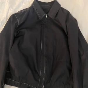 Svart jacka från Yohji Yamamoto - Säljer en stilren svart jacka från Yohji Yamamoto. Jackan har en dragkedja framtill och kontrasterande vita sömmar som ger en unik touch. Tillverkad i 100% bomull, vilket gör den både bekväm och hållbar. Perfekt för dig som vill ha något stilrent och tidlöst i garderoben. Priset är inte fast