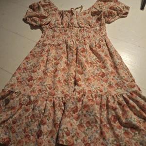 Blommig klänning med volangdetaljer - Charmig blommig klänning med volangdetaljer och korta puffärmar. Den har en smockad midja och knytning i ryggen för en söt look. Perfekt för sommarens alla tillfällen!