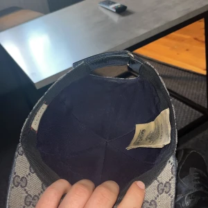 Grå mönstrad keps med justerbart spänne - Snygg grå keps med ett ikoniskt mönster och justerbart spänne baktill. Kepsen har en randig detalj i rött och blått på sidan. Perfekt för en stilren look.