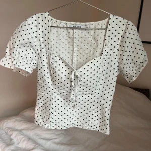 Vit prickig blus från NA-KD - Säljer en söt vit blus med svarta prickar från NA-KD. Blusen har korta ärmar och en knytning framtill som ger en fin detalj. Perfekt för en somrig look! 🌼
