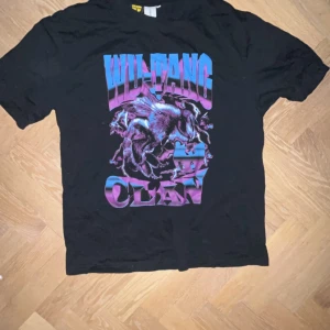 Wu-Tang Clan t-shirt från Junkyard - Säljer en svart t-shirt från Junkyard med ett coolt Wu-Tang Clan-tryck i blått och lila. Perfekt för fans av hiphop och streetwear. T-shirten har korta ärmar och en klassisk passform. Skön och stilren för alla tillfällen.