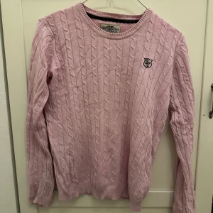 Rosa stickad tröja från Hampton Republic - Säljer en söt rosa stickad tröja från Hampton Republic med kabelstickat mönster och ett broderat emblem på bröstet. Tröjan har långa ärmar och rund halsringning. Perfekt för kyliga dagar!💕 Svarar så snabbt så möjligt🤗