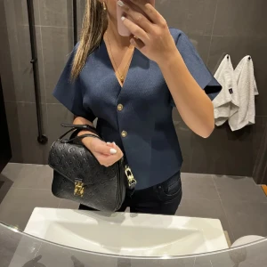 Blå Zara topp  - Snygg blå topp från Zara med korta ärmar och v-ringning. Den har två dekorativa knappar framtill och en stilren design som passar perfekt för en chic look. Helt oanvänd med prislapp. Storlek S🥰