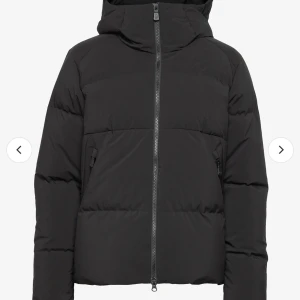 Svart dunjacka från Sail Racing - Snygg svart dunjacka från Sail Racing med huva och dragkedja. Jackan har en stilren design med logotypdetaljer på ärmen och bröstet. Perfekt för kalla dagar.