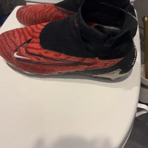 Röda och svarta fotbollsskor från Nike - Snygga fotbollsskor från Nike i rött och svart med ett iögonfallande mönster. Skorna har en strumpliknande passform och är perfekta för fotbollsplanen. De har en tydlig Nike-logga på sidan.